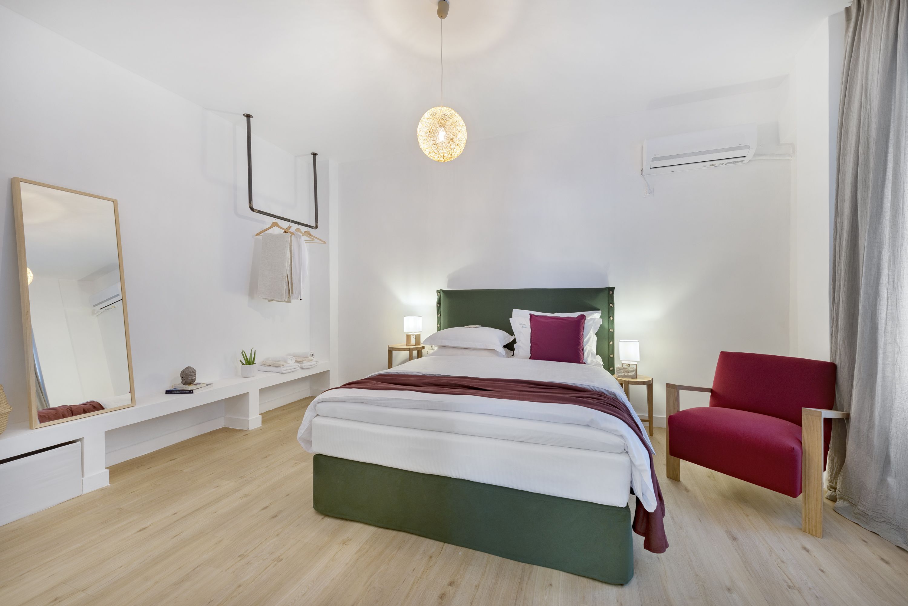 A modern bedroom with a bed, a red chair, a hanging light fixture, and a wall-mounted air conditioner.