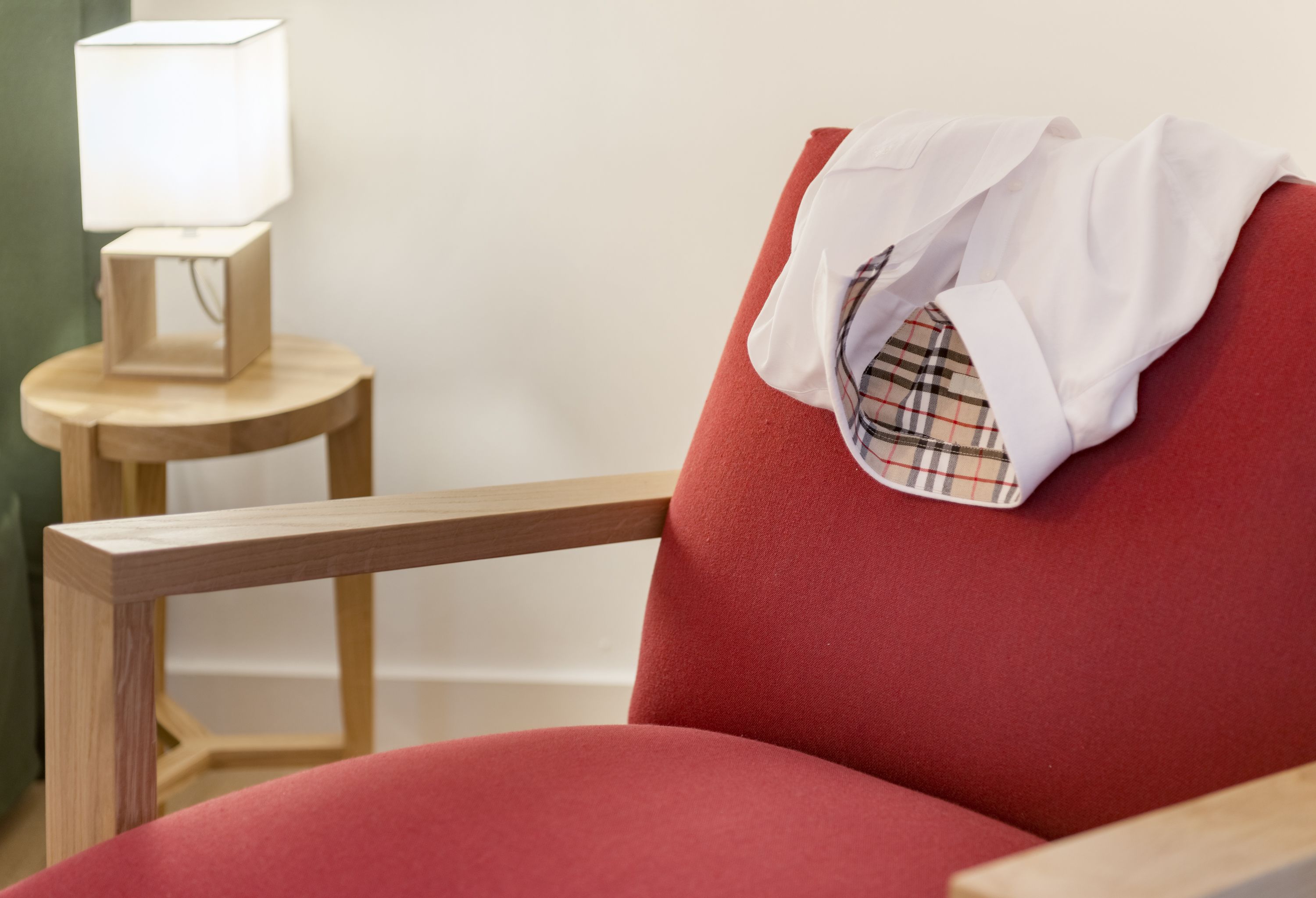 Une chaise rouge, une chemise à carreaux jetée dessus, une petite table en bois avec une lampe, et un mur sobre en arrière-plan.