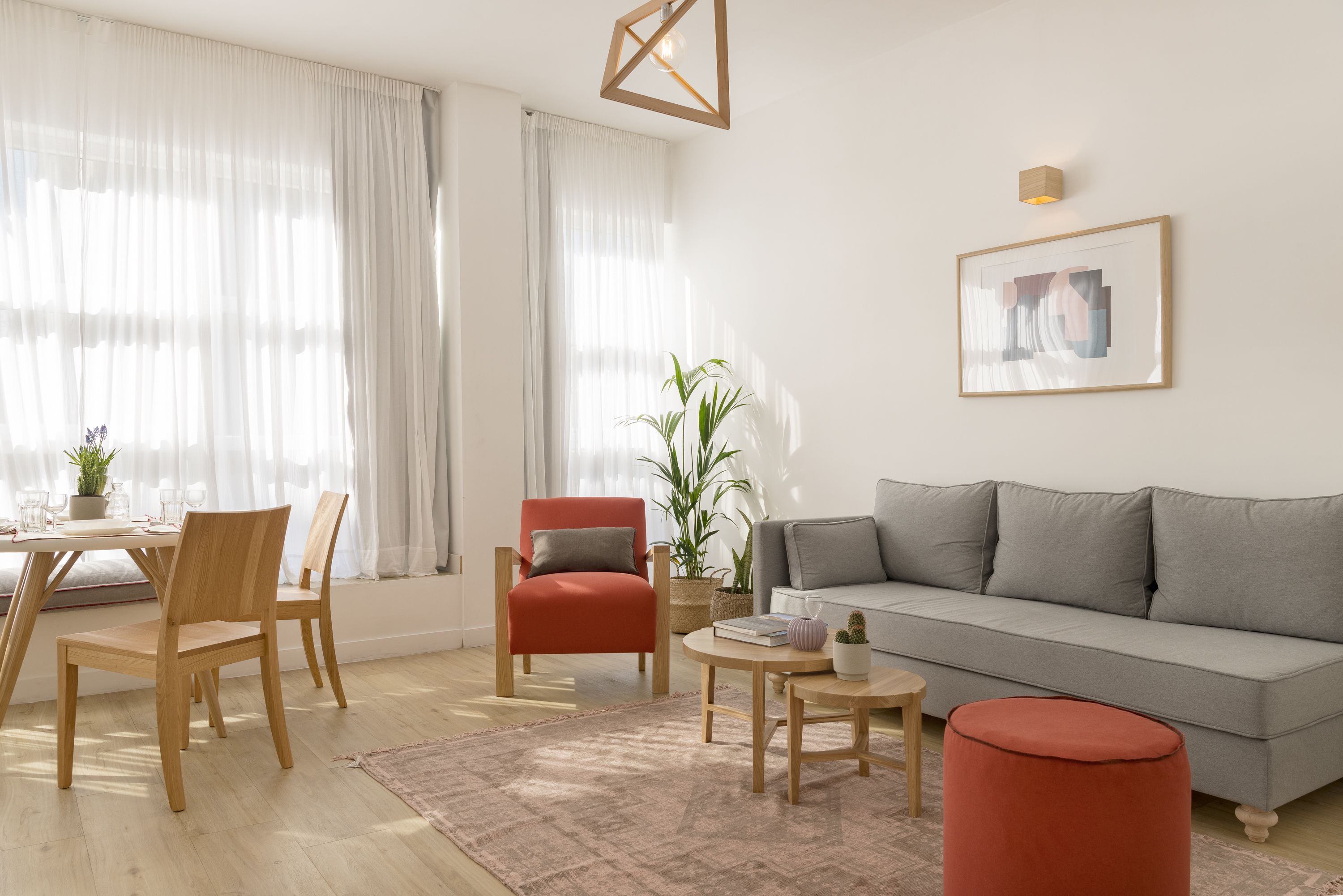 Een moderne woonkamer met een grijze bank, houten stoelen, een rode fauteuil, een salontafel en een eettafel met stoelen.