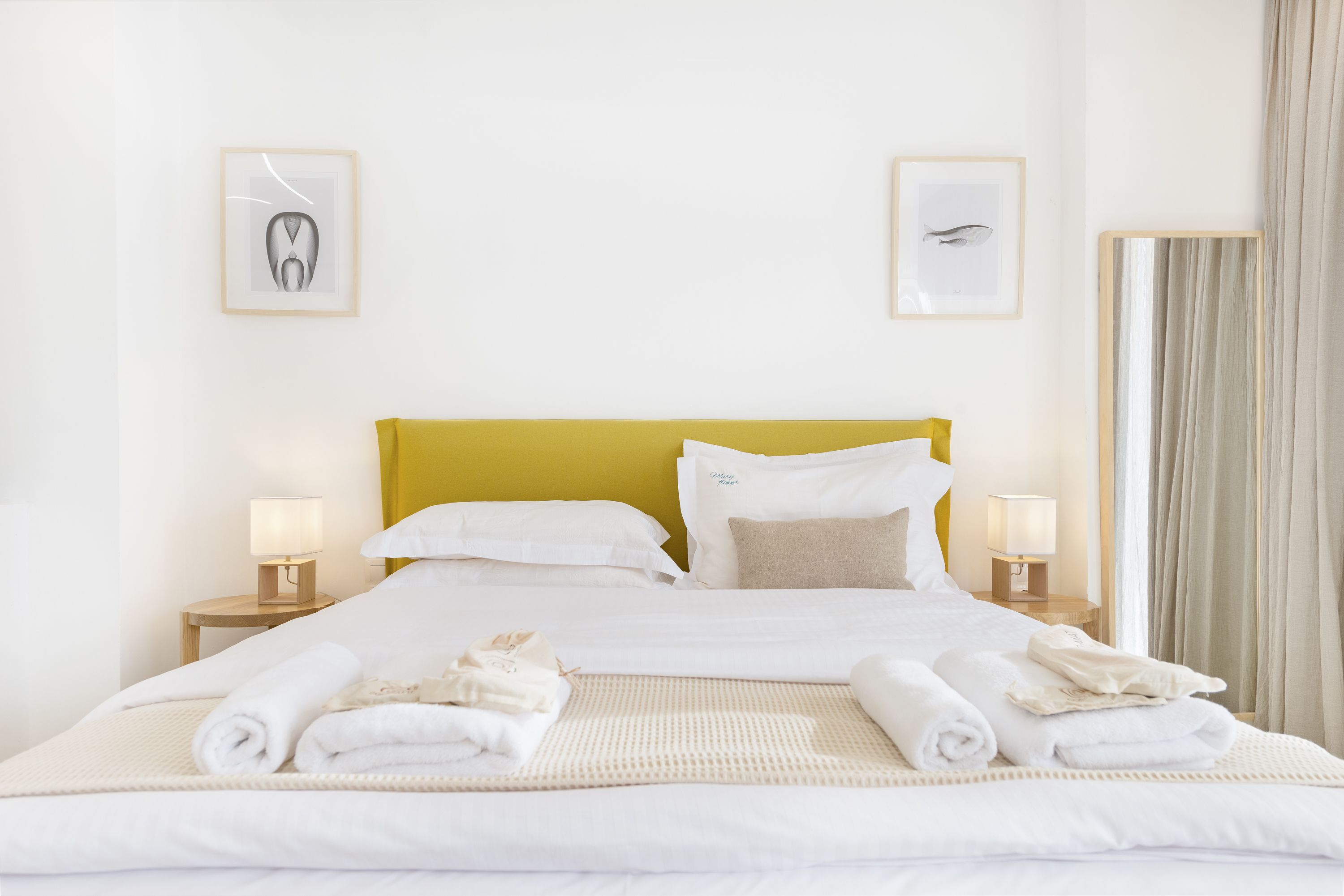A neatly made bed with white linens and rolled towels, a yellow headboard, and two bedside lamps.