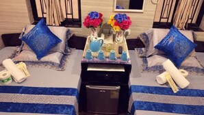 Habitación de hotel con dos camas, cada una con colcha azul y blanca a cuadros, y almohadas azules a juego. Hay una mesita con un florero de flores coloridas, una tetera, tazas y un control remoto. Se ve un refrigerador al frente.