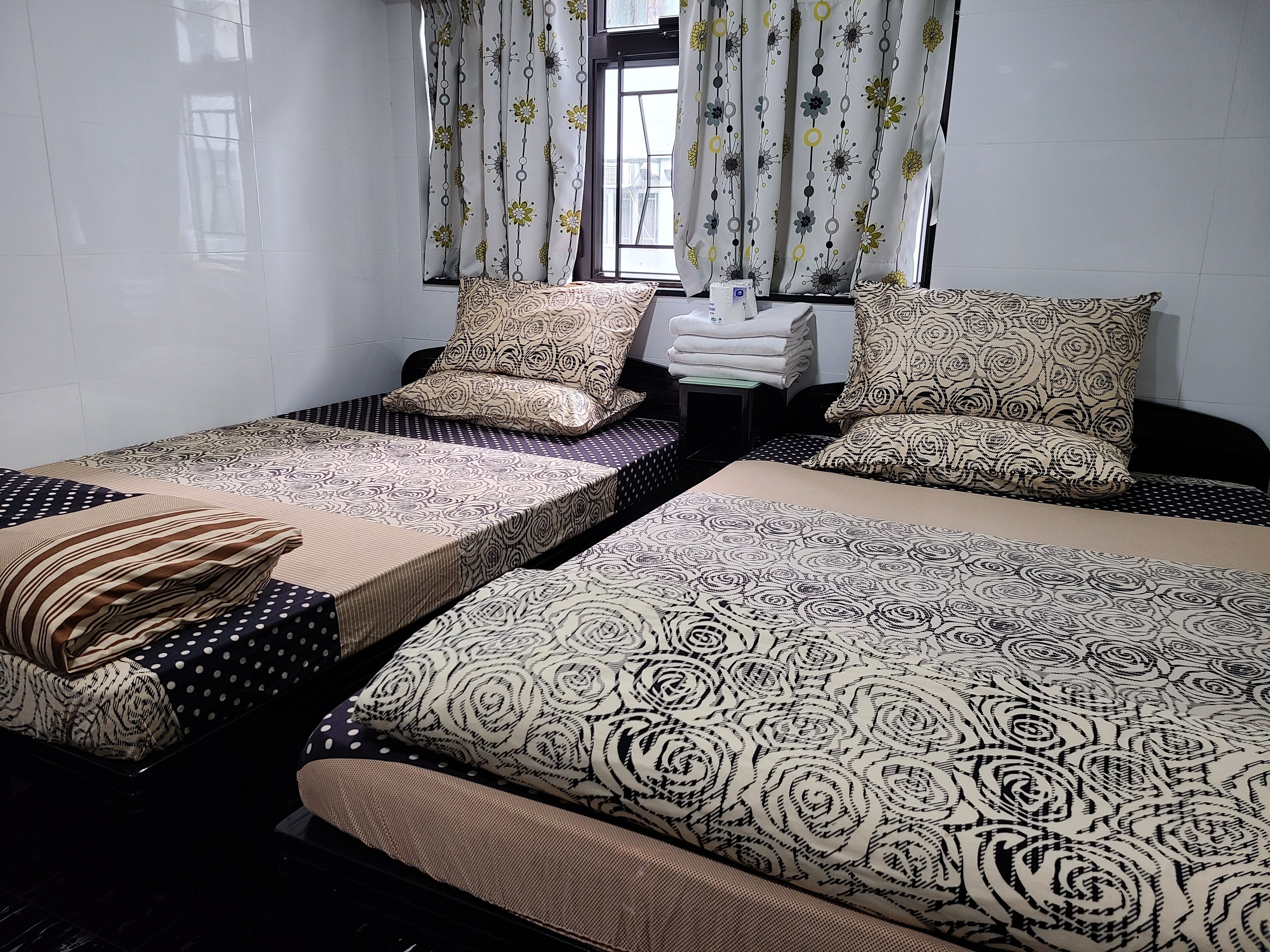 Una habitación con dos camas individuales, cada una con ropa de cama y almohadas estampadas. Se ve una ventana con cortinas florales, una mesita con toallas y una silla.