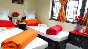 Habitación de hotel con dos camas, cada una con ropa de cama blanca y roja, almohadas naranjas y una mesita con un florero de flores rojas.