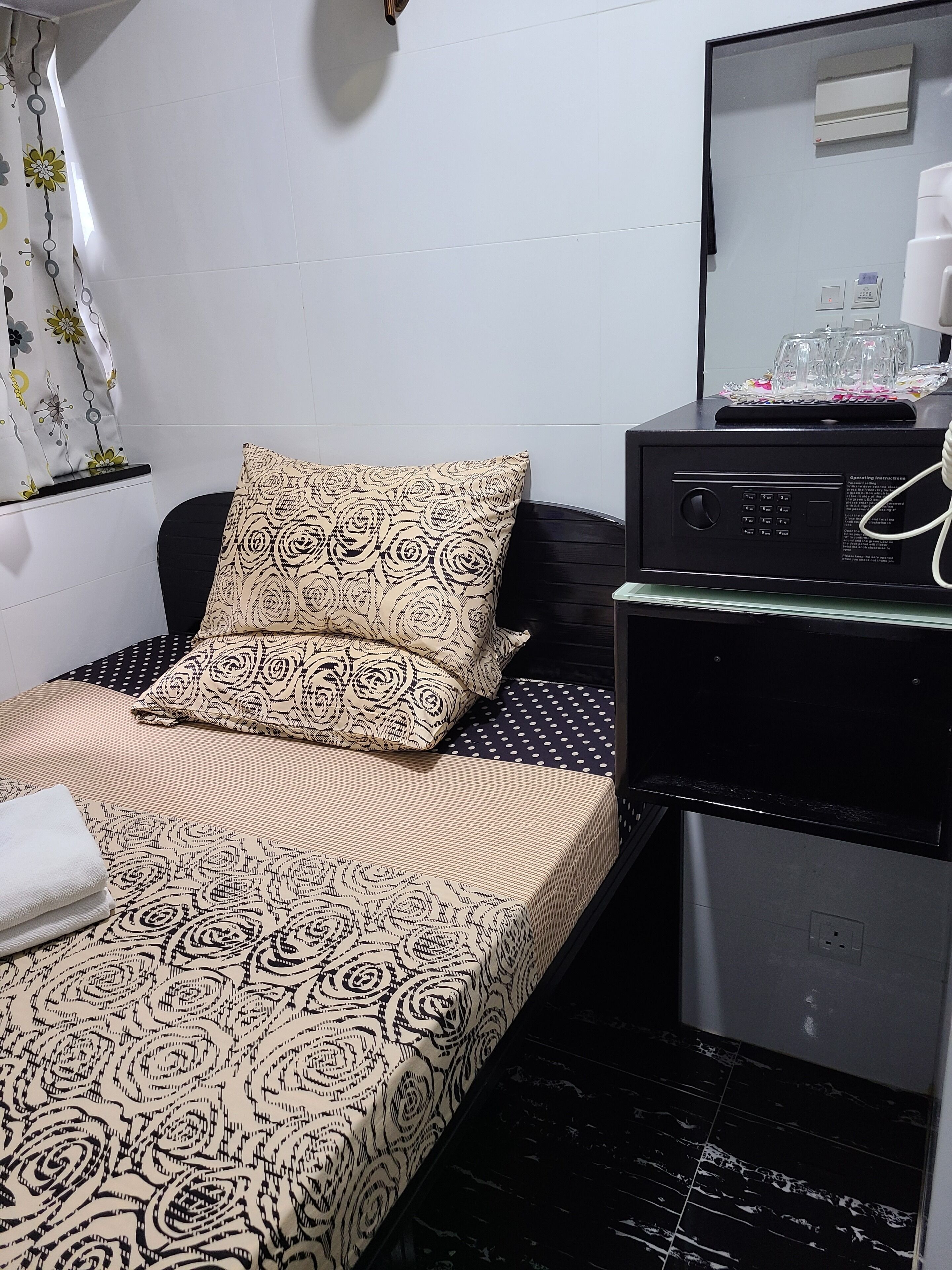 A single bed with patterned bedding, a black headboard, a small bedside table with a mirror, and a black suitcase.
