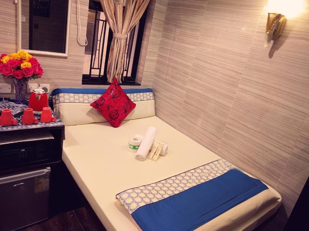 A compact hotel room with a bed, a small table with toiletries, a vase of flowers, and a window with curtains.