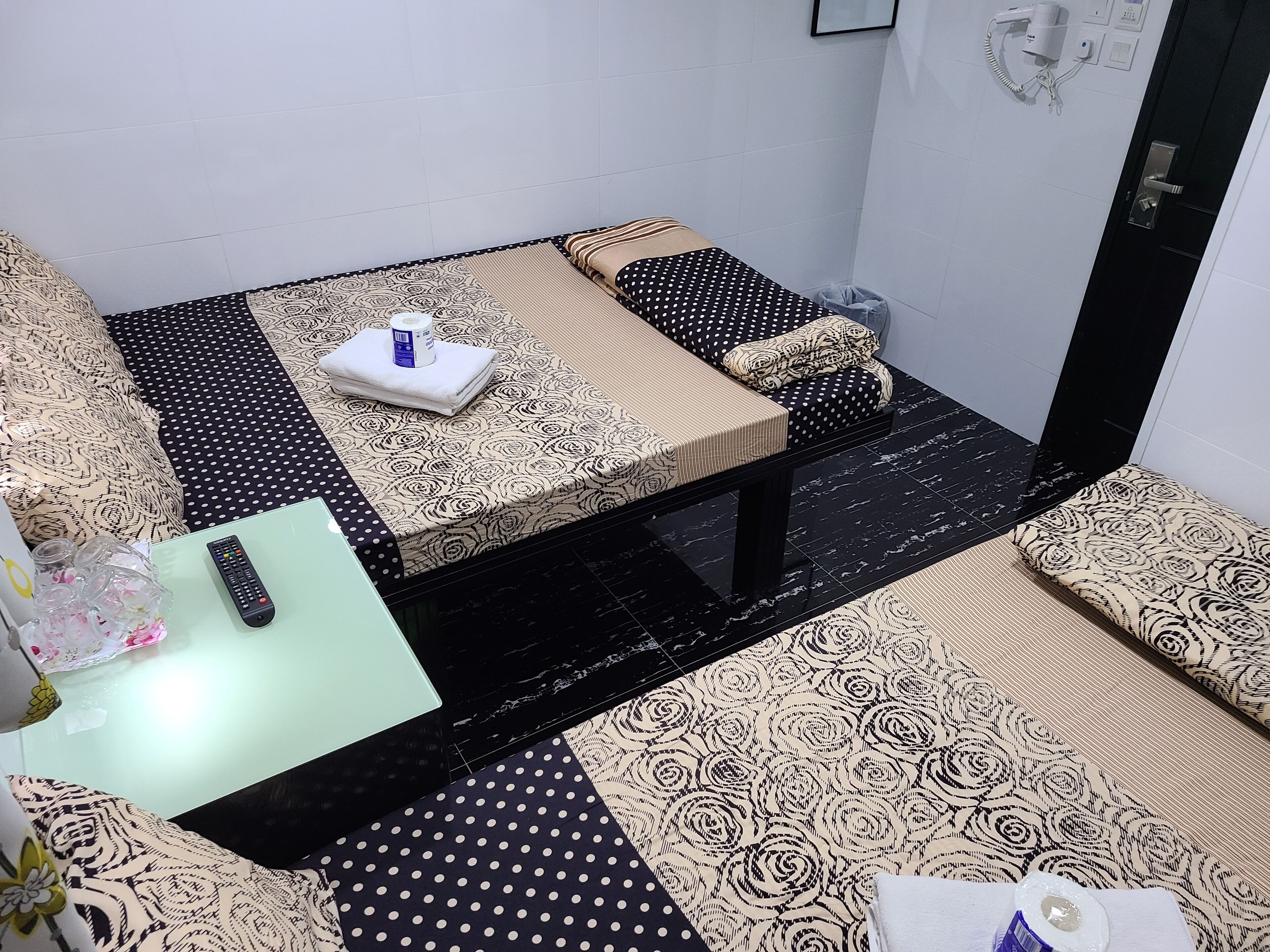 A compact room with a bed, a small table, and a chair. There is a hairdryer on the wall and a trash bin near the door.