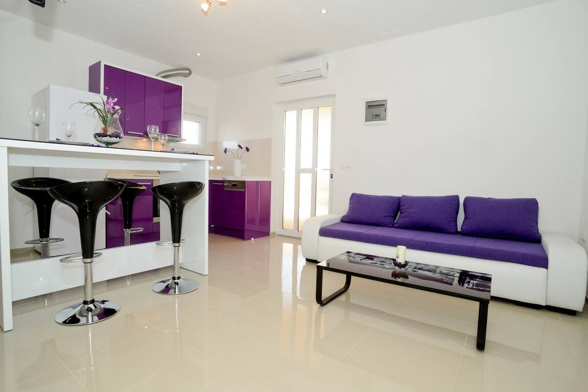 A modern living room with a white sofa and purple cushions, a glass coffee table, and two black bar stools.
