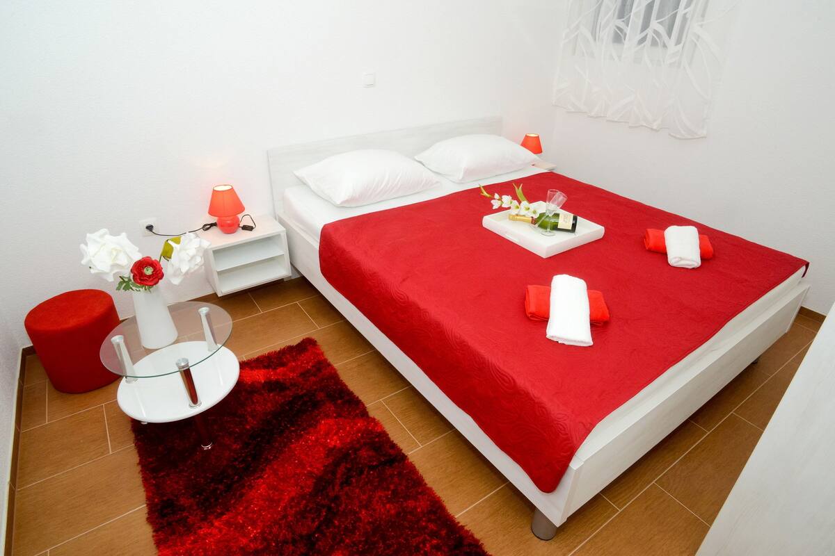 A modern hotel room with a large bed, red bedding, a glass side table, a red ottoman, and a red rug.