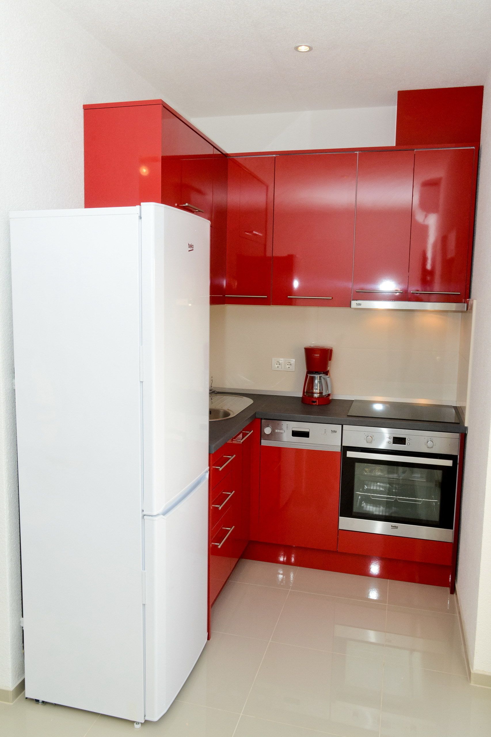 A compact kitchen with red cabinets, a white refrigerator, a coffee maker, and a built-in oven.