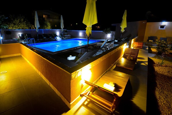 An outdoor swimming pool area with lounge chairs and umbrellas, surrounded by a low wall and a well-lit deck. - Apartments Julijana (Privlaka)