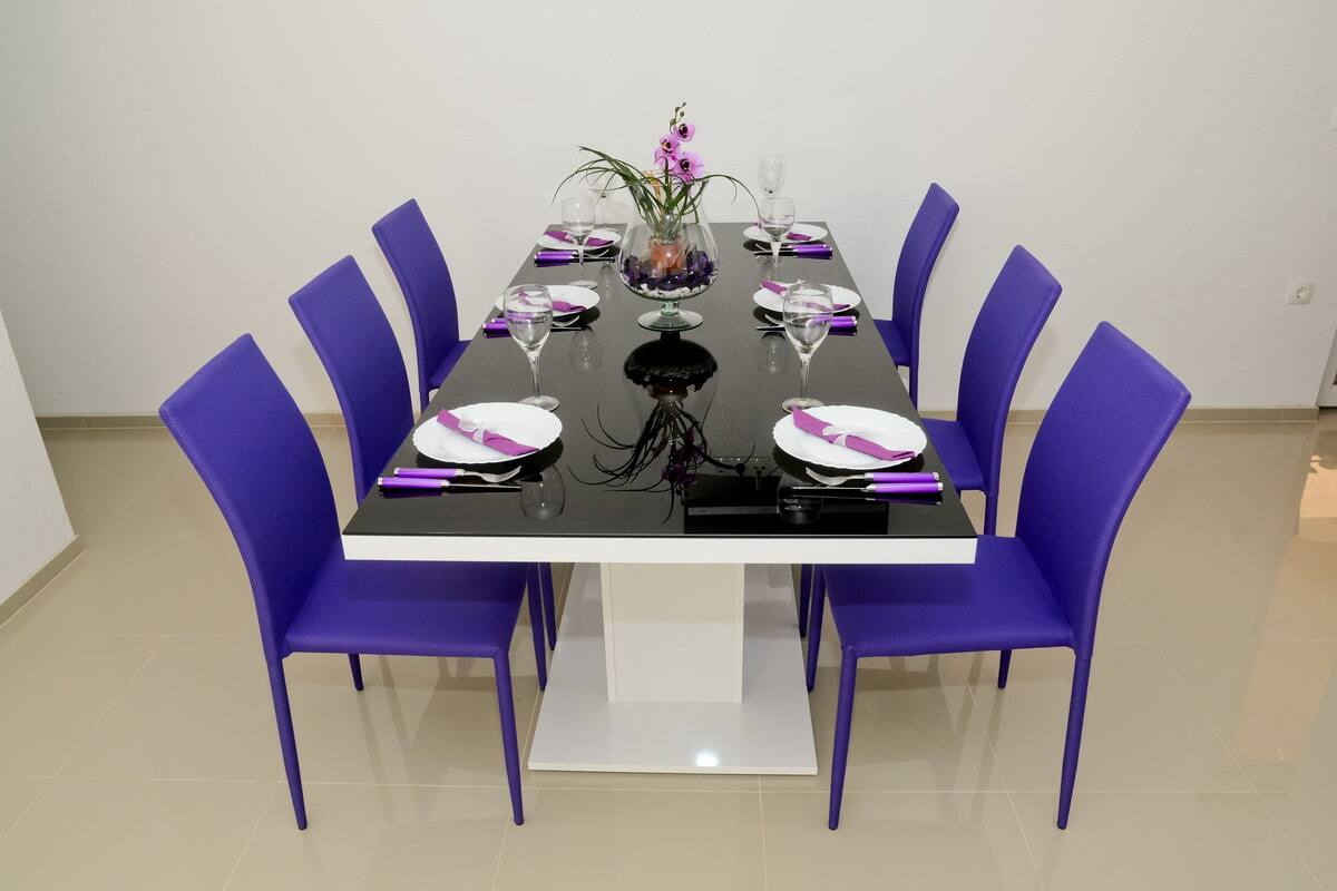 A dining table with purple chairs, a vase with flowers, and wine glasses set for six.