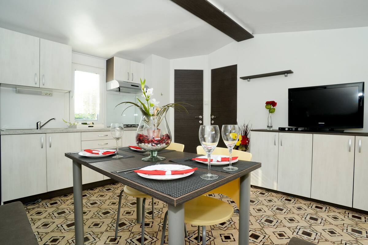 A modern kitchen with white cabinets, a dining table set with plates and glasses, and a flat-screen TV mounted on the wall.
