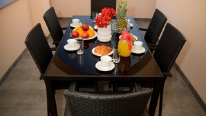 Mesa de comedor lista para cuatro personas, con sillas, vista a edificios a través de un arco, y una mesa con frutas y un jarrón de flores.