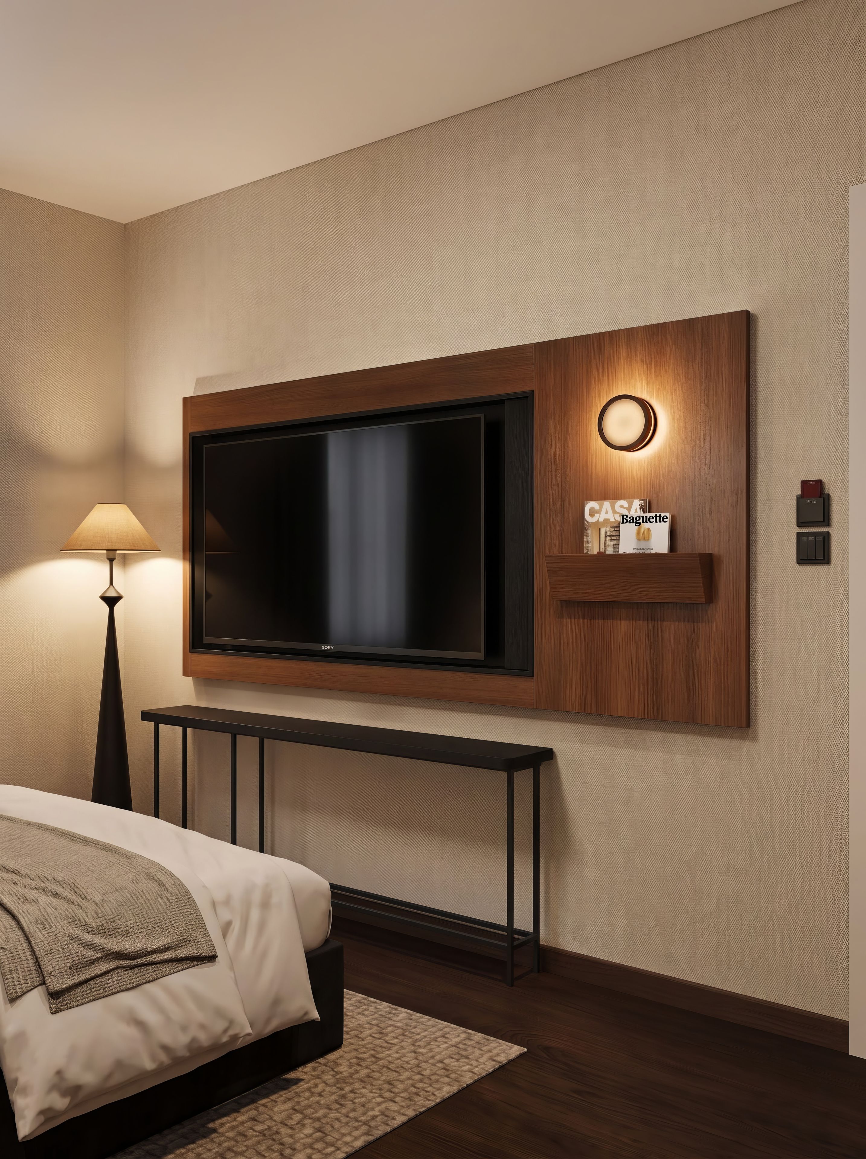 A modern hotel room with a large flat-screen TV mounted on a wooden panel, a bedside table, a lamp, and a magazine rack.