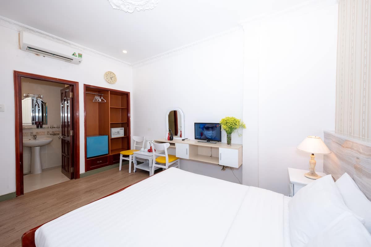 A hotel room with a bed, a desk with a chair, a TV, a bedside table with a lamp, a wardrobe, and a bathroom with a sink and mirror.