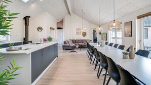 Eine moderne Küche mit einer zentralen Kochinsel, einem Essbereich mit langem Tisch und Stühlen sowie einem Wohnbereich mit Sofa und Couchtisch.