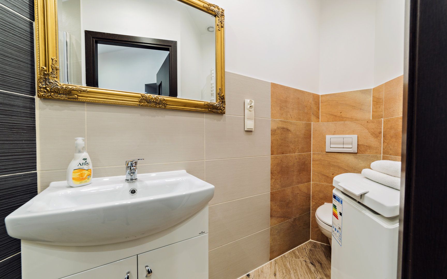 Un baño con un lavamanos blanco, un espejo con marco dorado y un inodoro con un adhesivo de arcoíris.