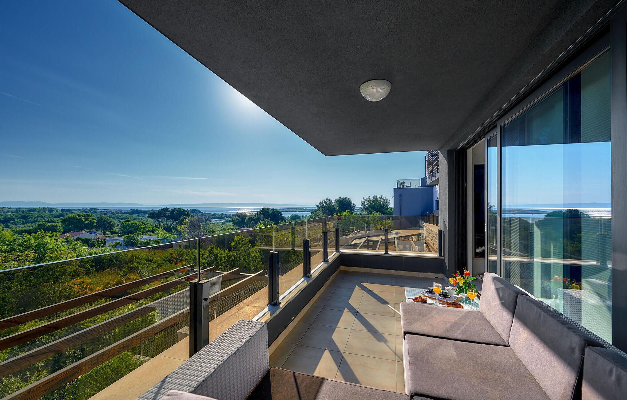 Ein Balkon mit Blick auf eine Küstenlandschaft, ein Glastisch mit Kissen und moderne Architektur.