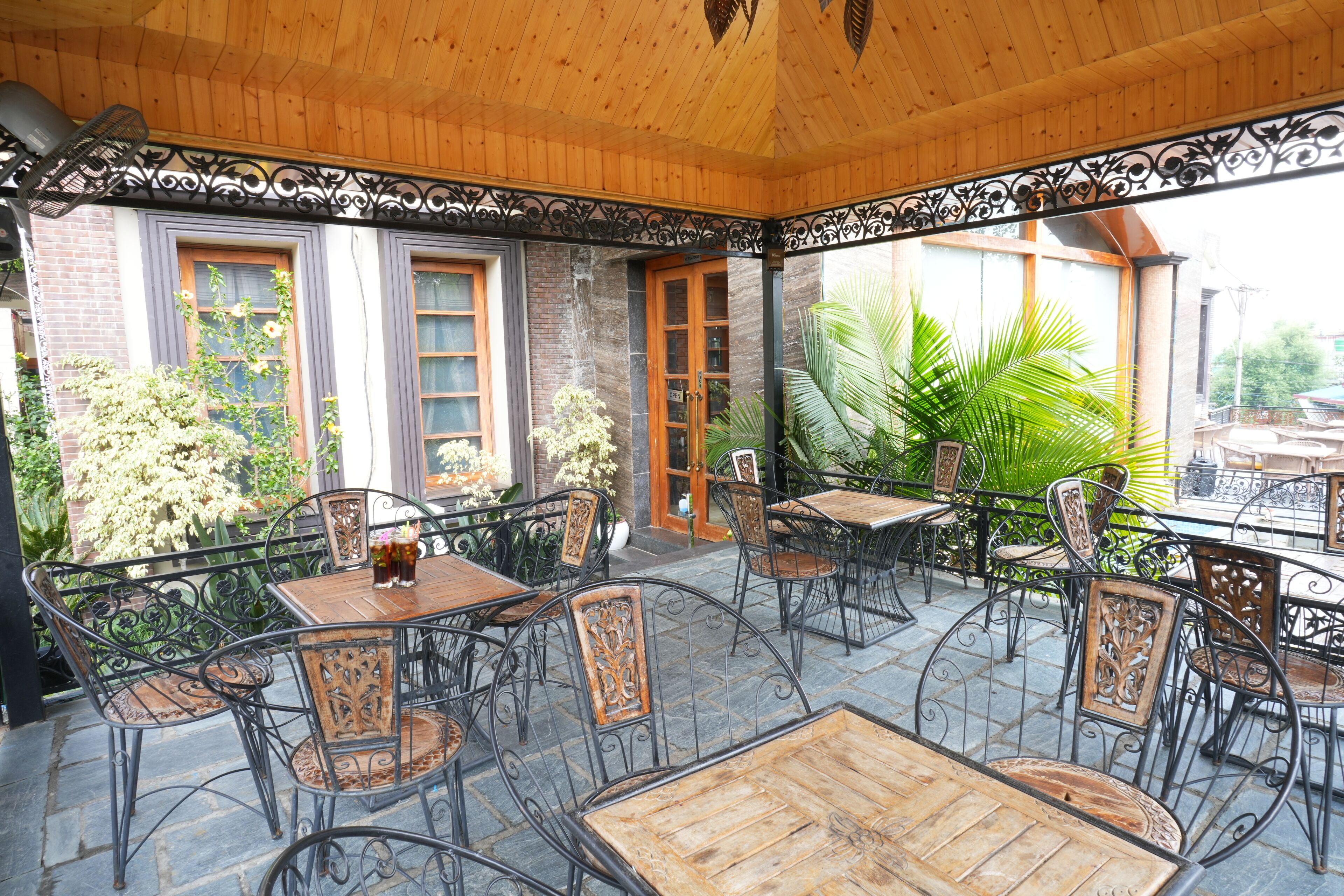An outdoor patio with wooden tables and metal chairs, surrounded by brick walls and large windows.
