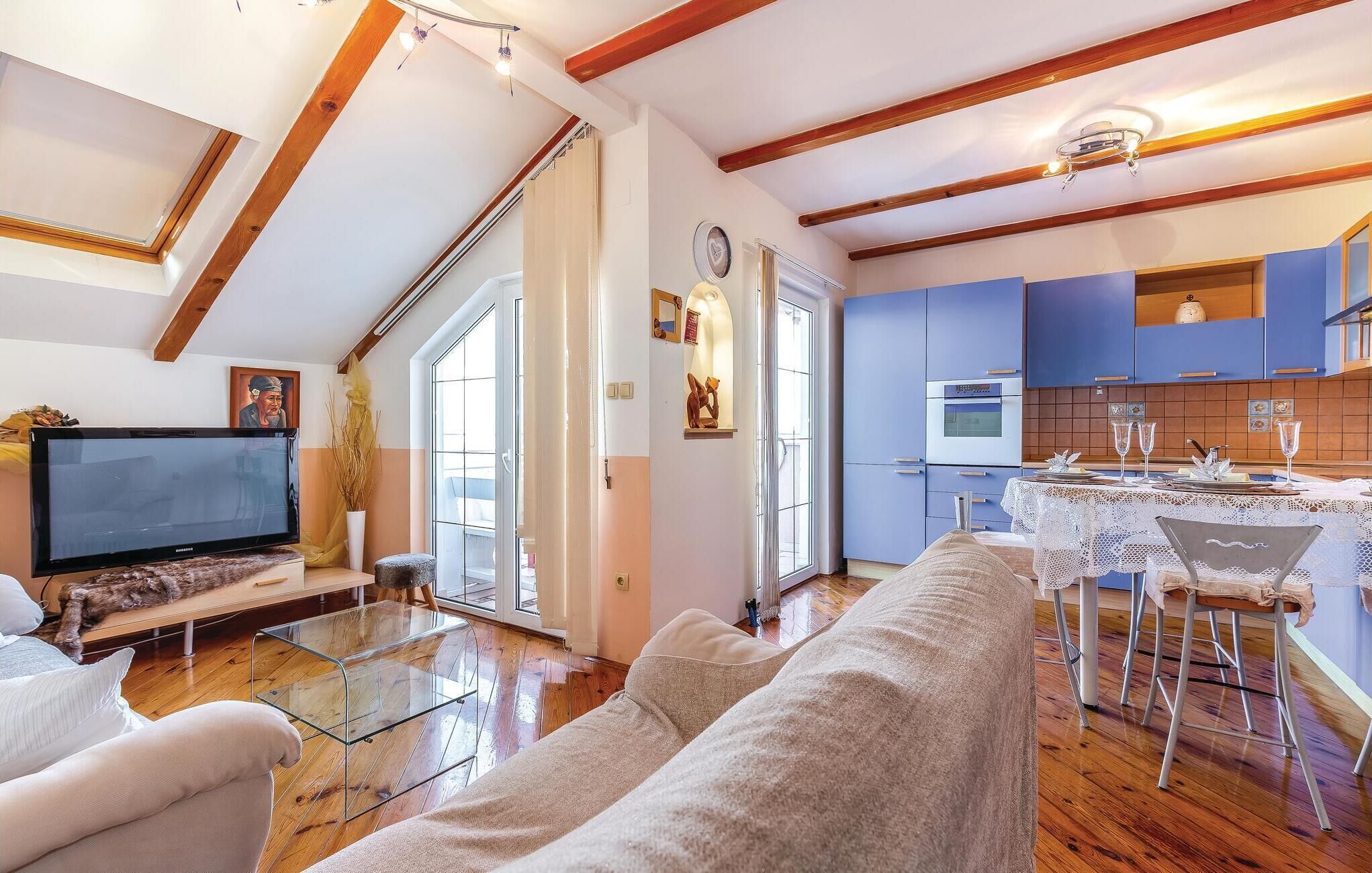 Uma sala de estar moderna com uma TV de tela plana, uma mesa de centro de vidro e um sofá bege. Há uma área de cozinha com armários azuis e uma mesa de jantar para dois.