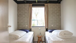Una habitación pequeña con dos camas individuales, un radiador, una ventana con cortinas y un taburete de madera.
