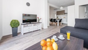 Una sala de estar moderna con una televisión de pantalla plana, una mesa de centro con frutas y bebidas, un sofá gris y un área de comedor al fondo.