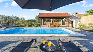Una piscina con sillas de descanso y sombrillas, un área de estar cubierta y un edificio de piedra al fondo.