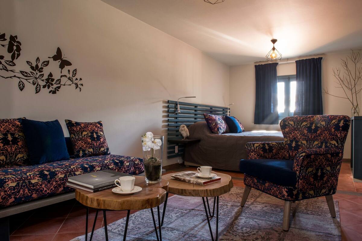 Een gezellige woonkamer met een bank, fauteuil en salontafel, een bed met een blauw hoofdeinde, en een raam met gordijnen.