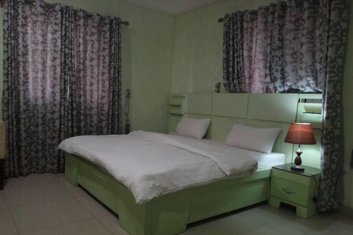 Un dormitorio con cama, mesita de noche, lámpara y cortinas.