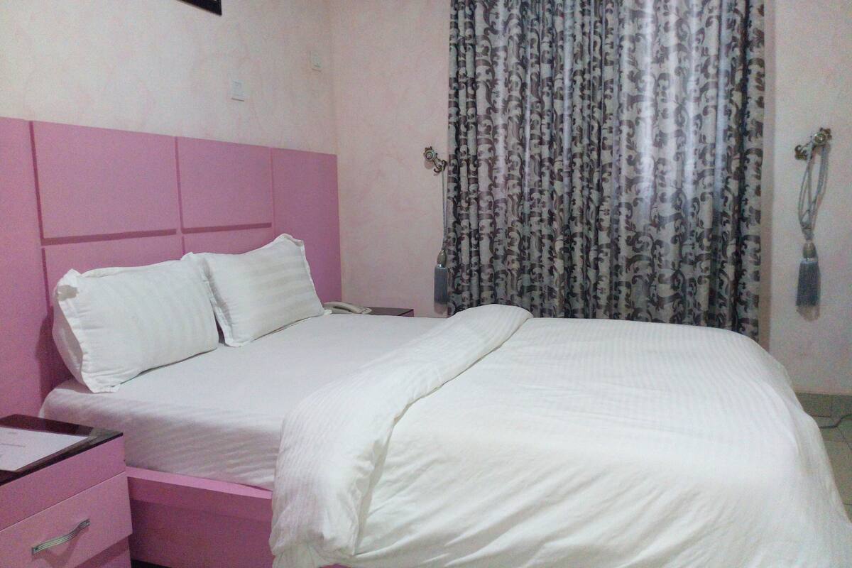 Un dormitorio rosa con cama, mesita de noche y cortina.