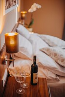 Una cama bien hecha con sábanas blancas, una botella de vino y dos copas de vino vacías sobre una mesita de noche de madera.