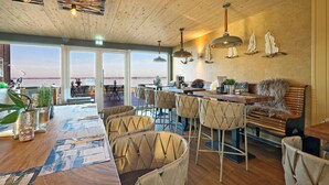 A dining area with a wooden ceiling, large windows overlooking a body of water, and a bar with stools. - Beautiful apartment in Lembruch/Dümmer See with indoor swimming pool (Lembruch/Dümmer See)