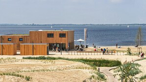 A modern wooden building with a glass facade, located near a sandy beach with a view of the water and wind turbines in the distance. - Beautiful apartment in Lembruch/Dümmer See with indoor swimming pool (Lembruch/Dümmer See)