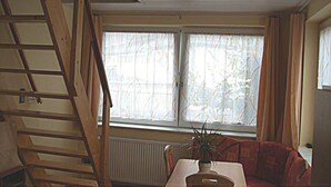Een kamer met een houten trap, een radiator, een raam met gordijnen en een tafel met een plant.