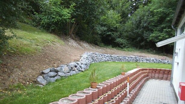 Een aangelegde tuin met een stenen keerwand, een geplaveid pad en een gazon met potplanten.