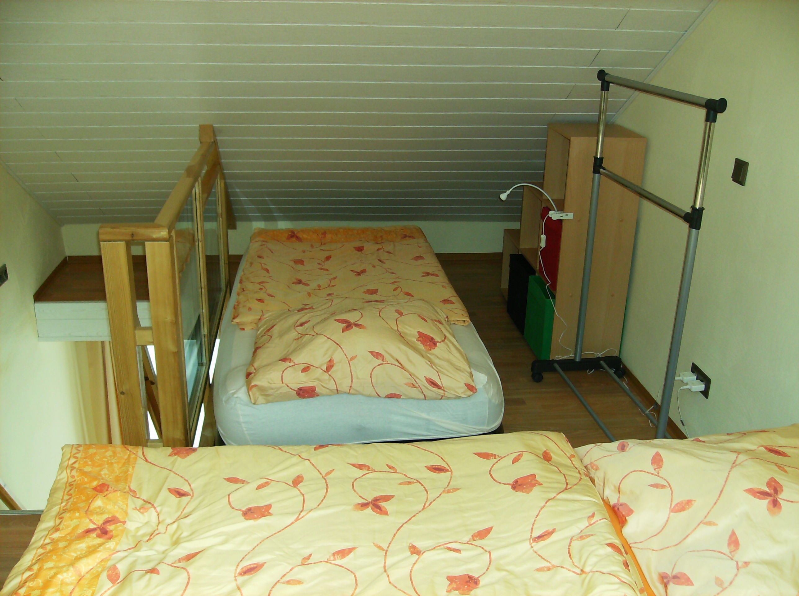 Una habitación pequeña con cama individual, una escalera de madera, un escritorio y un perchero de metal.