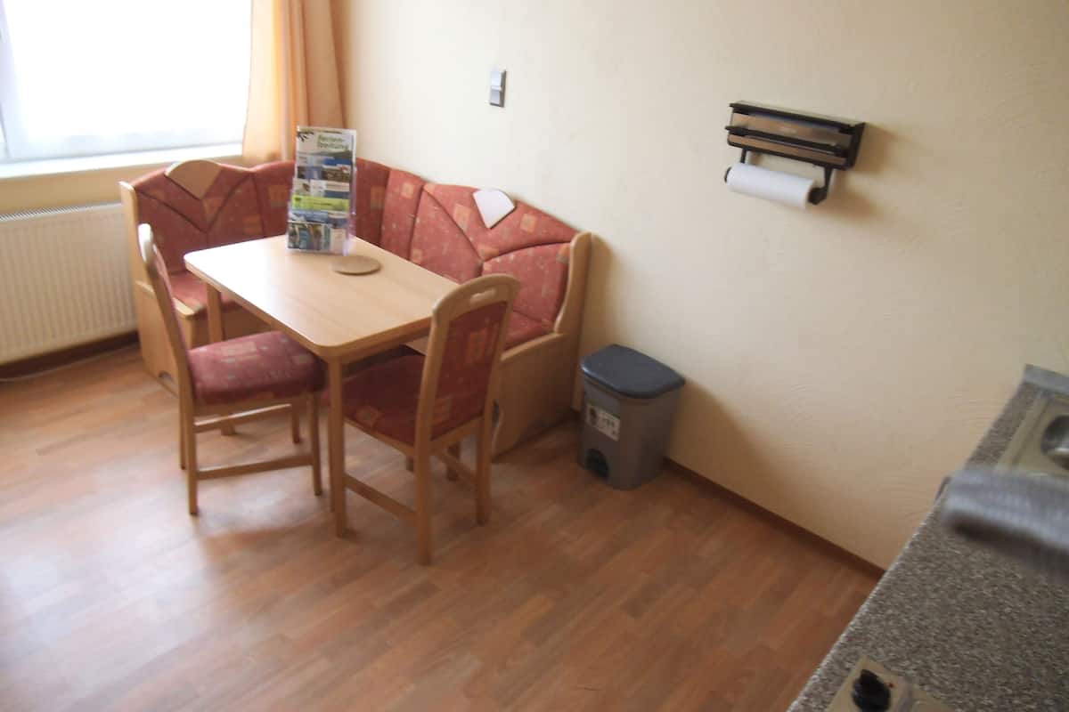 Une petite pièce avec une table en bois et deux chaises, un canapé rouge, un radiateur, une poubelle et un tableau d’affichage au mur.
