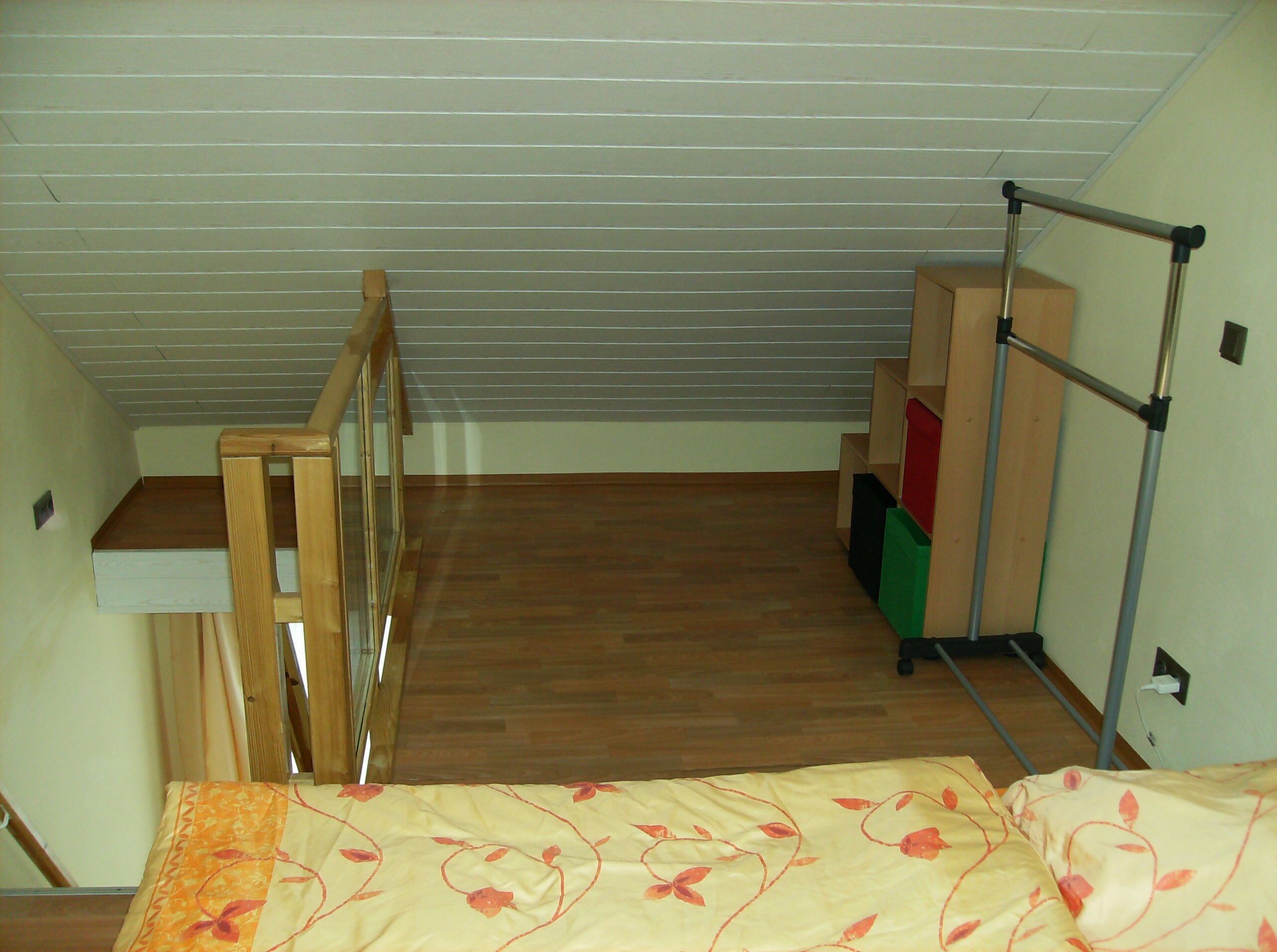 A small, narrow room with a sloped ceiling, a wooden bench, a bed with a floral bedspread, and a metal rack with storage boxes.