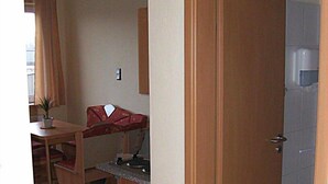 Een compacte kamer met een houten deur, een kleine kitchenette, een eethoek met tafel en stoelen, en een zichtbare prullenbak.