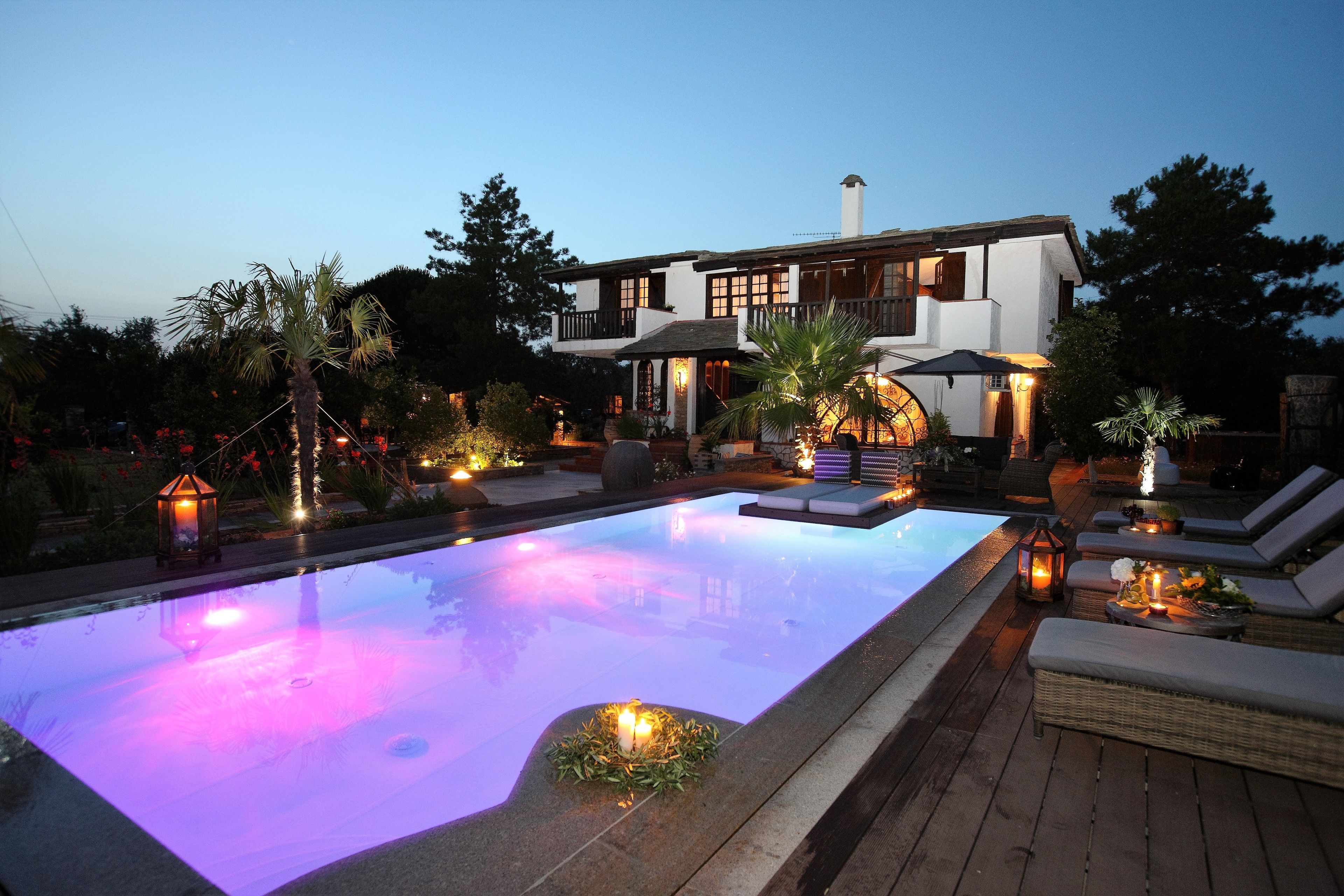 Piscina com deck de madeira, cadeiras de descanso e uma casa ao fundo.
