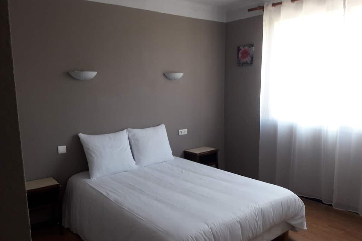 Una cama bien hecha con sábanas blancas en una habitación con piso de madera, pared marrón y ventana con cortinas de gasa.