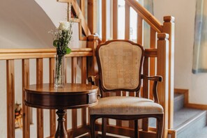 A wooden chair with a cushioned seat and backrest, a round wooden table with a vase of flowers, and a wooden staircase in the background. - Gästehaus Rösch (Wiesent)