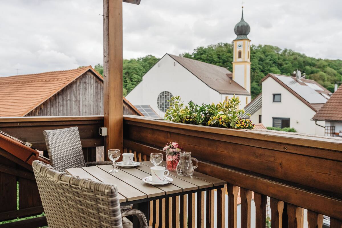 A wooden balcony with wicker chairs and a table set for dining, overlooking a church and residential buildings.