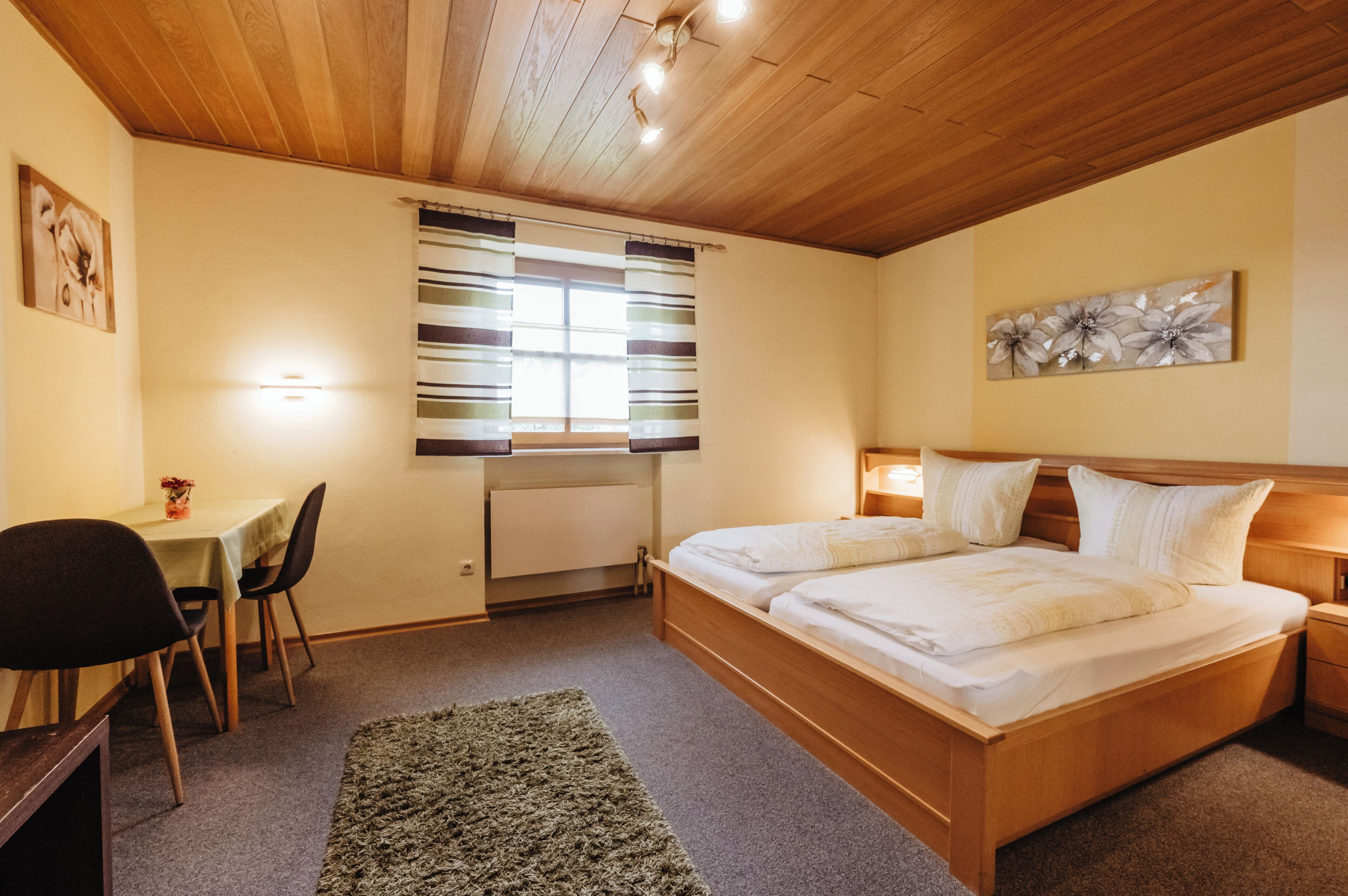 Un dormitorio con techo de madera, una cama con ropa blanca, un escritorio con silla y una ventana con cortinas a rayas.