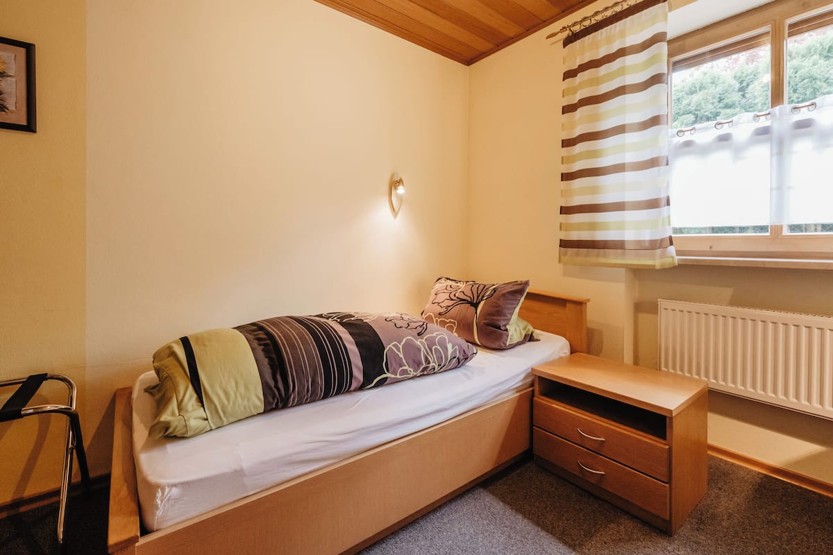 A single bed with a headboard, a bedside table with drawers, a wall-mounted light, a radiator, and a window with striped curtains.