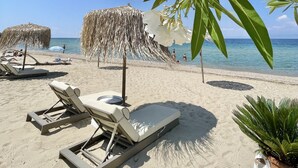 A beach with lounge chairs, a thatched umbrella, and white flowers. - Byblos Mare (Thasos)