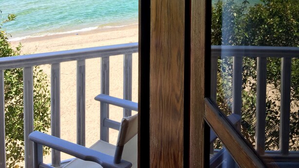 A view from a balcony with a wooden railing, overlooking a beach with clear turquoise water and a sandy shore. - Byblos Mare (Thasos)