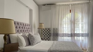 A bedroom with a tufted headboard, a bedside table with a lamp, a bed with patterned bedding, and sheer curtains over a window. - Byblos Mare (Thasos)