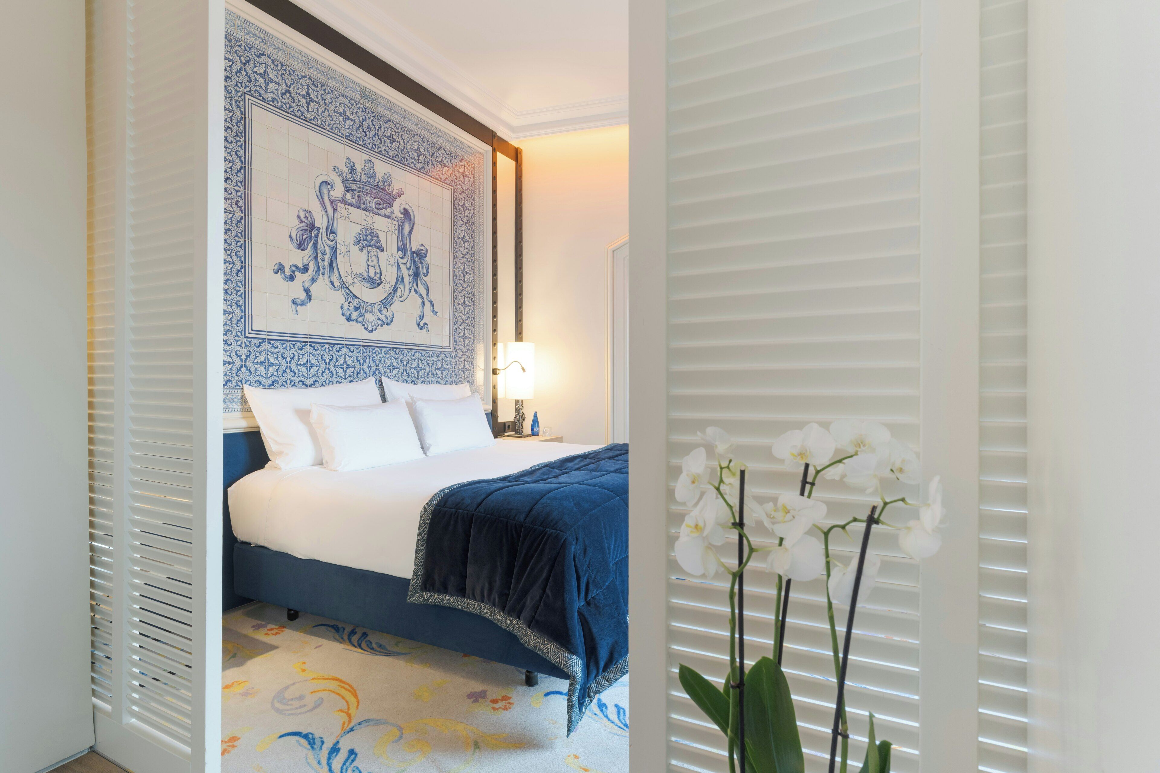 A hotel room with a large bed, a decorative wall tapestry, a nightstand with a lamp, and white orchid flowers.
