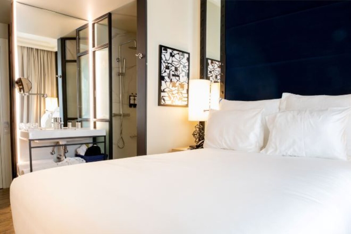 Una camera d'albergo con un letto grande, un bagno con uno specchio e un lavandino, e un lampadario fissato alla parete.